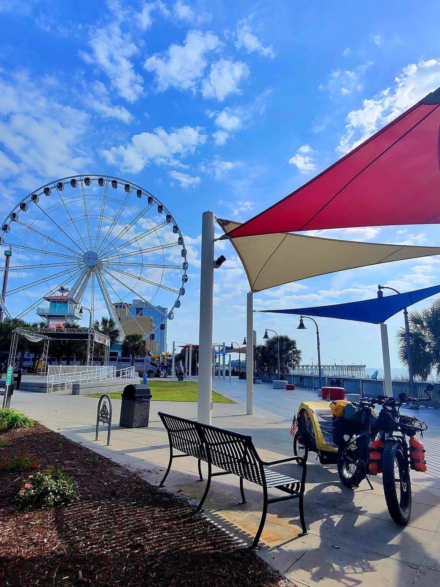 Real photo — enhanced: The Beast, Coach RODA's loaded e-bike, and The Sherpa, his Burley trailer, on the Myrtle Beach boardwalk under the shade sails, the SkyWheel behind — Apr 22, 2025